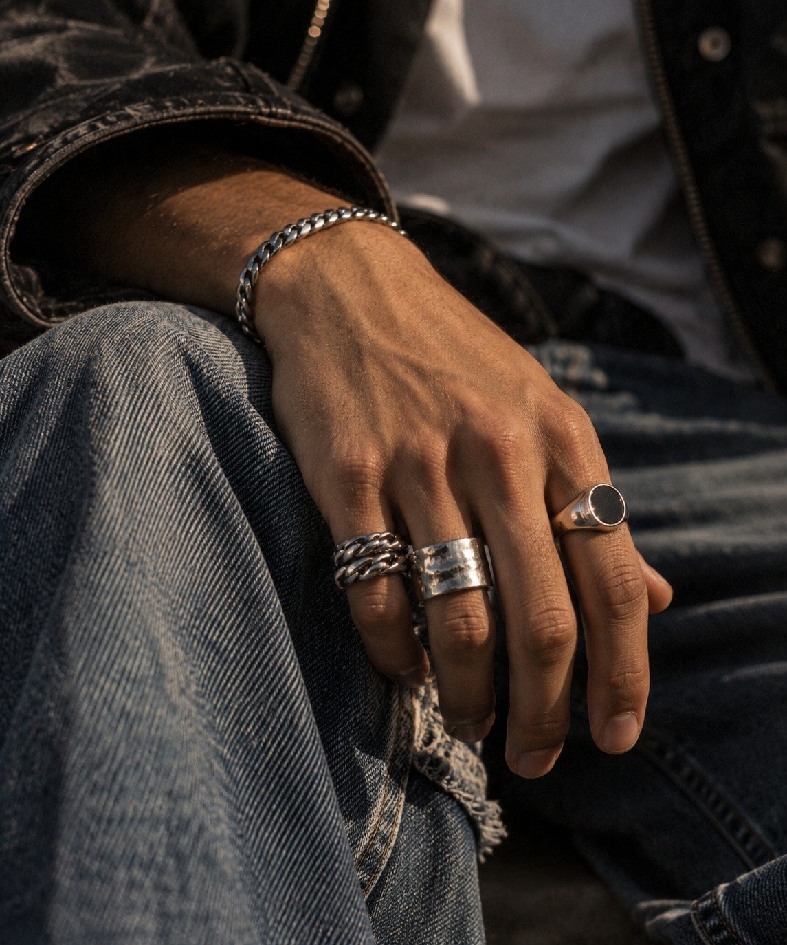 Hand with stacked silver rings and an onyx signet resting on denim